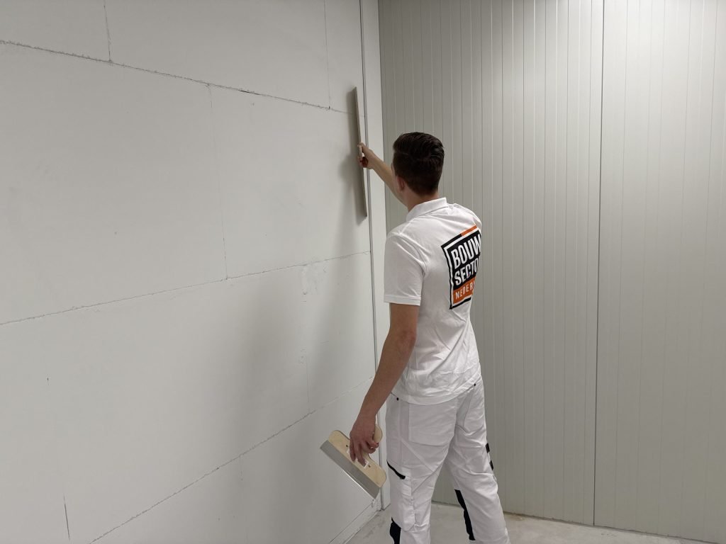 Stukadoor in wit werkpak loopt met de spaan langs de rand van een wand bij een lichte deur