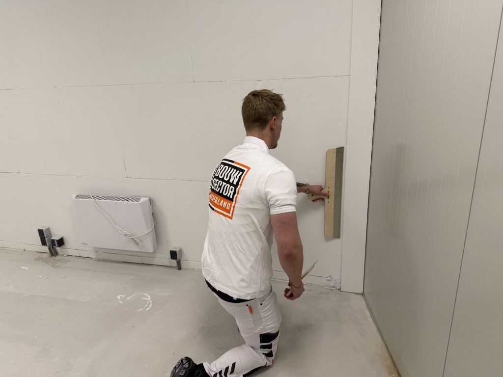 Stukadoor zit geknield bij een kachel en werkt de onderkant van een muur strak af