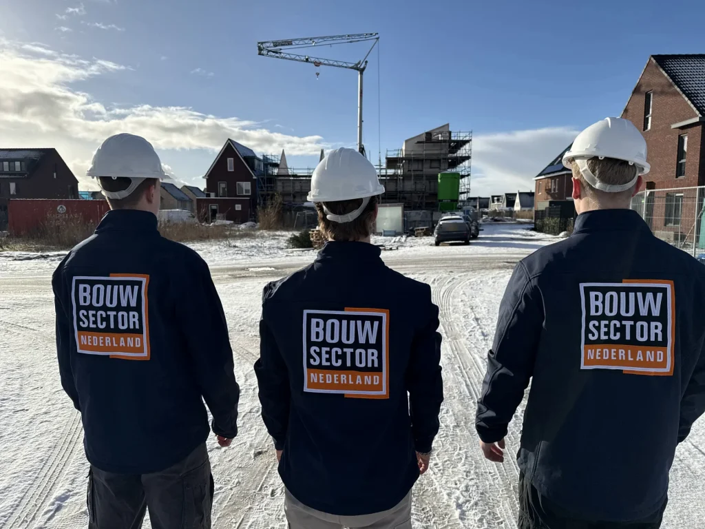 Drie vakmensen in donkerblauwe jassen van Bouwsector Nederland kijken naar een nieuwbouwwijk in de sneeuw